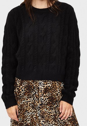 Schwarzer Strickpullover mit Kabelstruktur und gerippten Bündchen, kombiniert mit einem Rock im Leopardenmuster, der vorne mit Knöpfen versehen ist.