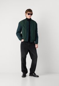 Jachetă bomber verde cu textură matlasată, pulover negru cu guler înalt, blugi negri largi și pantofi negri; accesorizată cu ochelari de soare închiși.