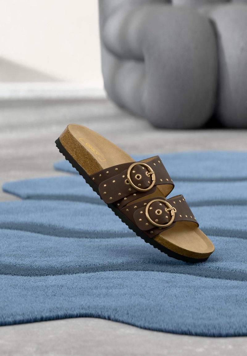 Sandale slide marron avec deux boucles dorées et des clous, semelle en liège, posée sur un tapis bleu texturé avec un poivron flou en arrière-plan.