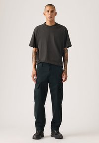 Μαύρο γκρι μπλουζάκι με κοντά μανίκια, συνδυασμένο με σκούρο παντελόνι cargo με πολλαπλές τσέπες και χαλαρή γραμμή. Μαύρα παπούτσια ολοκληρώνουν την εμφάνιση.