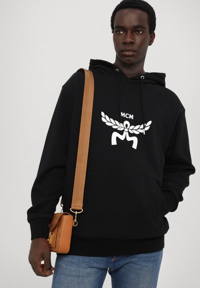 Hoodie noir avec logo MCM blanc sur la poitrine. Présente des cordons de serrage et une poche kangourou. Associé à un sac bandoulière marron et à un jean bleu.