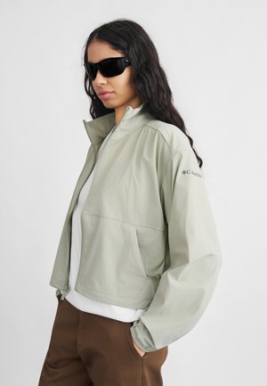 Vrouw met lang donker haar, zwarte zonnebril, lichtgrijs jasje met rits, wit shirt en een bruine broek met handen in de zakken.