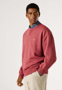 Sudadera de algodón roja con puños y cuello de canalé, que presenta un pequeño logo blanco. Llevada sobre una camisa de botones azul y pantalones beige con textura.