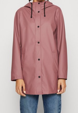 Person iført en dæmpet lyserød hættetrøje med sorte trykknapper og lommer, kombineret med blå jeans.