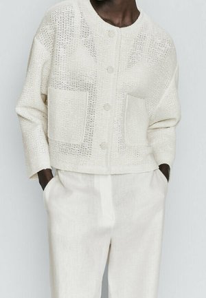 Modèle portant un cardigan en maille crème boutonné avec deux poches avant et un pantalon ample taille haute assorti. Mains dans les poches.