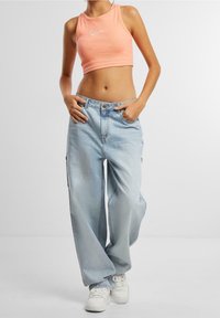 Mladá osoba oblečená v bezramenném broskvovém crop topu, volných světle modrých džínách a bílých teniskách, stojící s rukama v kapsách před jednobarevným pozadím.