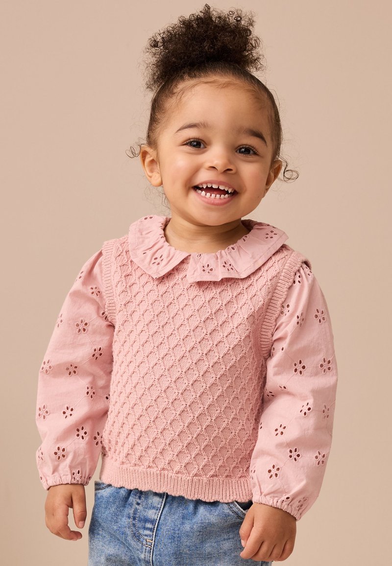 Pull rose en maille avec un motif texturé en forme de losange, associé à de longues manches roses ornées de broderies florales. Un short en denim complète le look.