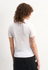 T-shirt branca de mangas curtas com um design ajustado e gola canelada, feita de tecido leve. As costas apresentam uma tatuagem visível.