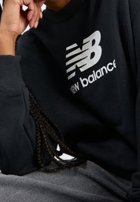 Felpa nera con un grande logo bianco di New Balance, con polsini a costine e scollatura a girocollo. Tessuto testurizzato con una vestibilità comoda.