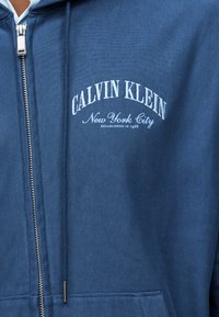 Zils rāvējslēdzēja džemperis ar baltiem izšuvumiem kreisajā krūšu daļā ar tekstu "Calvin Klein New York City Established 1968".