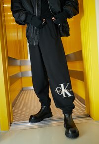 Svarta joggerbyxor med elastiska muddar, med en vit Calvin Klein Jeans-logotyp. Bärs med svarta klumpiga stövlar, mot en gul bakgrund.