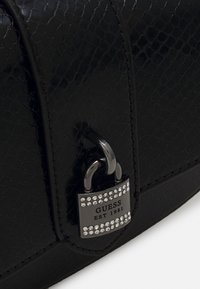 Sac en cuir de serpent noir embossé avec quincaillerie métallique, arborant une plaque ornée de diamants portant l'inscription "GUESS EST 1981." Texture lisse.