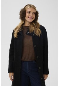 Donna con paraorecchie marroni, cappotto scuro testurizzato, maglione marrone e jeans blu, sorridente e che guarda di lato contro uno sfondo neutro.