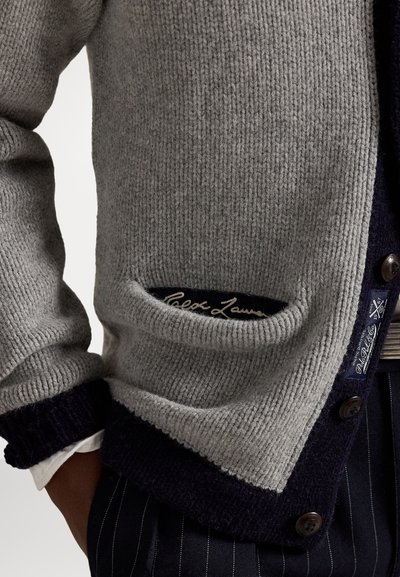 Sivi pleteni kardigan s mornarskim rubovima, gumbima i džepom na kojem se vidi vezena oznaka Ralph Lauren, nosi se preko prugastih hlača.