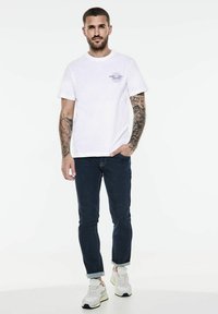 Bílý bavlněný tričko s malým grafickým detailem na hrudi, v kombinaci s tmavě modrými džíny a bílými teniskami. Standardní střih, krátké rukávy.