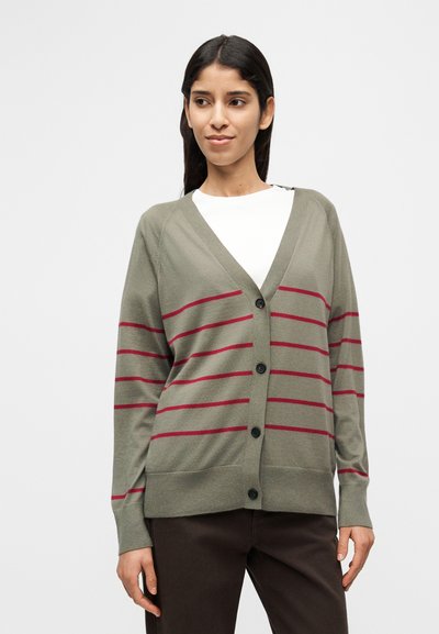 Πουλόβερ ελιάς με κόκκινες οριζόντιες ρίγες, V-λαιμόκοψη, κούμπωμα με κουμπιά, ριμπ μανσέτες και τελείωμα. Φορεμένο πάνω από ένα λευκό πουκάμισο, εμφανίζεται από μπροστά.