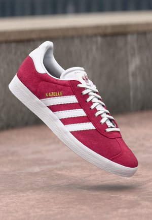 Rød Adidas Gazelle sneaker med hvide striber og snørebånd, hvid sål og guld "Gazelle" tekst, vist mod en udendørs urban baggrund.