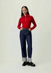 Un pull rouge en tricot avec un col zippé, associé à un jean bleu foncé taille haute avec un ourlet bleu clair. Des chaussures noires complètent le look.
