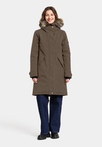 Brun parka med pelsbesat hætte, lynlåslukning, sidelommer og lange ærmer. Båret med vide blå jeans og sorte støvler.