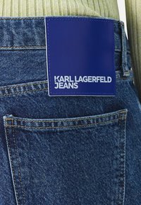 Denimové džíny s modrou koženou etiketou s nápisem "KARL LAGERFELD JEANS" bílým písmem. Tradiční pětikošová konstrukce a viditelné švy.