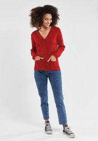 Rode gebreide cardigan met een V-hals, lange mouwen en knopen aan de voorkant; heeft twee zijzakken. Draagt met blauwe spijkerbroek en zwarte sneakers.