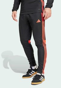 Pantalones deportivos negros con rayas naranjas, que presentan un corte ajustado, cintura elástica y detalle de logo, combinados con zapatillas negras y blancas.
