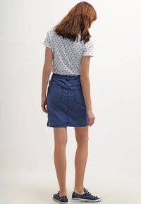 Mujer de pie con la espalda hacia la cámara, vistiendo una camisa blanca con un estampado, una falda de mezclilla azul y zapatillas de deporte en color navy.