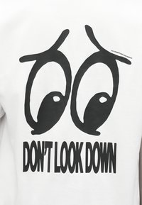 Μεγάλα στιλιζαρισμένα μαύρα μάτια και έντονο κείμενο «ΜΗΝ ΚΚΟΙΤΑΣ ΚΑΤΩ» τυπωμένα σε λευκό υφασμάτινο φόντο.