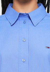 Világoskék, gombos ing gallérral, sima textúrával és egy kis logóval a bal oldalon, piros, fehér és fekete részletekkel.