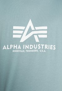 Camiseta de algodón azul claro que presenta un gran gráfico blanco con las letras "A" y "I" y texto que dice "ALPHA INDUSTRIES" y la ubicación.