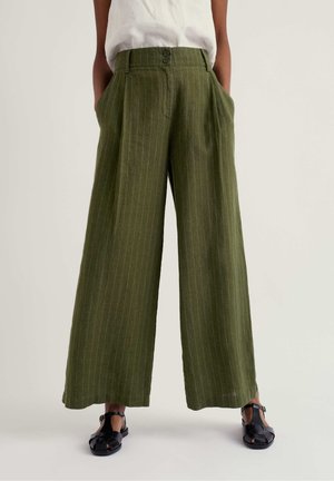 Model iført olivengrønne, højtaljede, viddebenede bukser med nålestriber og læg, kombineret med sorte sandaler med åben tå og en hvid top.