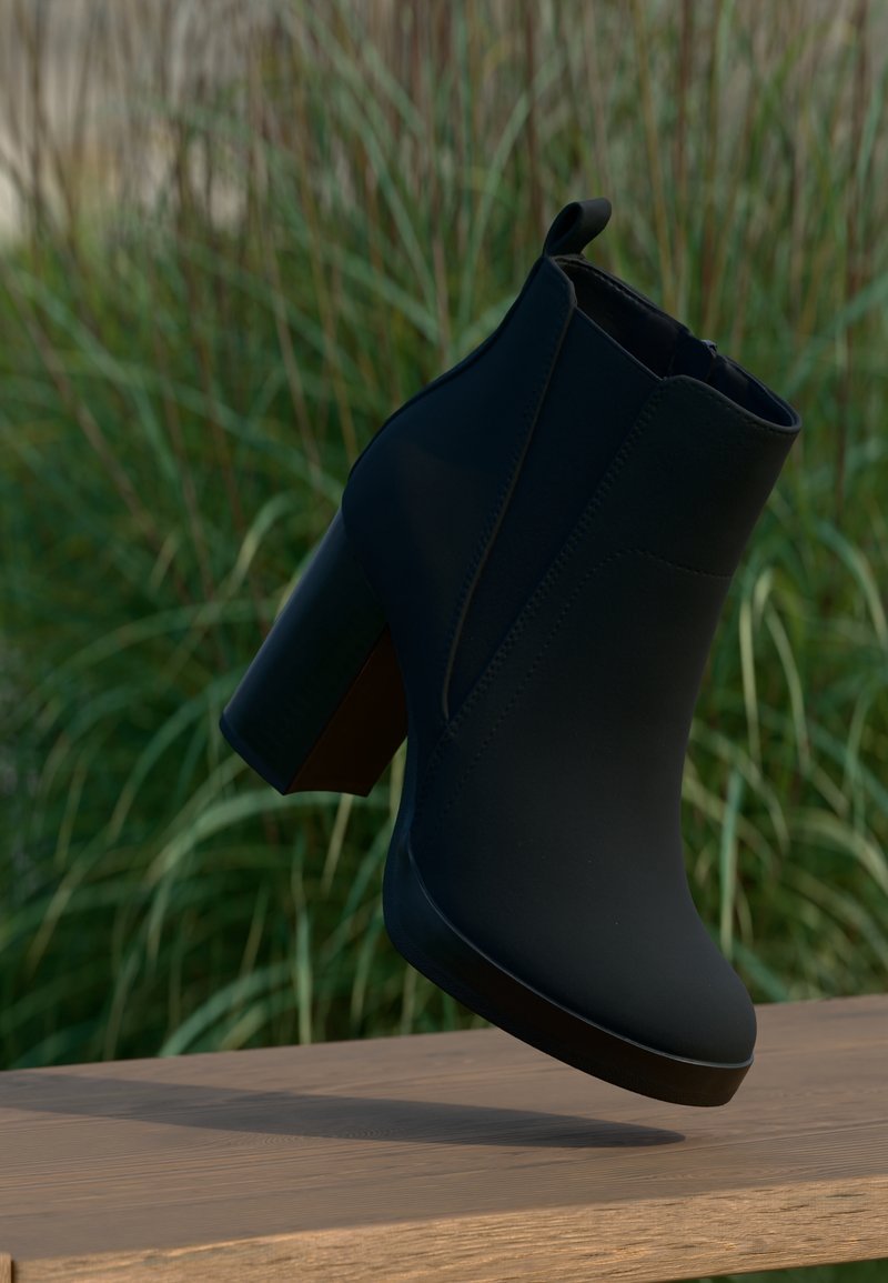 Bottine noire à talon carré posée au-dessus d'une surface en bois, avec en arrière-plan une herbe haute verte floue.