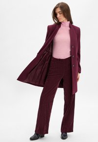 Bordeaux lange jas met vijf knopen, gedragen over een lichtroze coltrui en bijpassende corduroy wijde broek, die een rijke textuur laat zien.