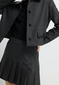 Femme portant une veste gris foncé à fines rayures avec des boutons noirs et une jupe plissée assortie, associée à un haut noir, posant avec la main sur la hanche.