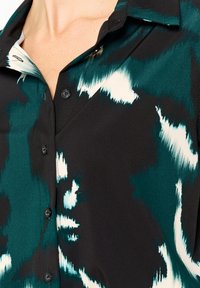 Short-sleeve shirt in black with green and white abstract pattern, featuring a pointed collar and black buttons along the front placket.