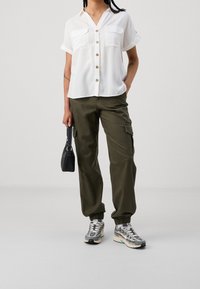 Camicia bianca a maniche corte con bottoni e due tasche, abbinata a pantaloni cargo verde oliva con polsini elasticizzati e una borsa nera. Le sneakers completano il look.