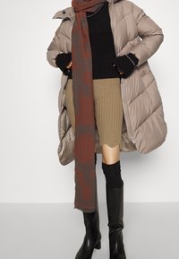 Kvinde iført beige puffjakke, sort sweater, brun ribbet nederdel, lang brun og grå tørklæde, og sorte knæhøje støvler, står med hænderne på hofterne.