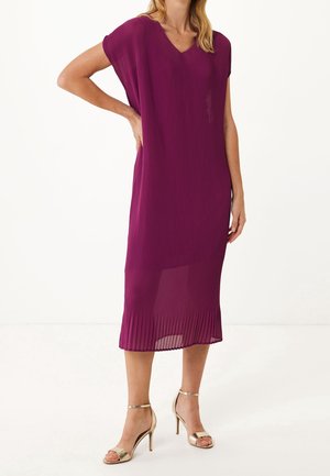 Femme portant une robe violette sans manches, longueur genou, plissée, avec un décolleté en V et des sandales à talons hauts dorés, debout avec une main sur la hanche.