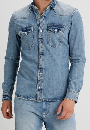 Mężczyzna w jasnoniebieskiej dżinsowej koszuli na guziki z dwoma kieszeniami na piersi oraz dopasowanych jasnoniebieskich dżinsach.