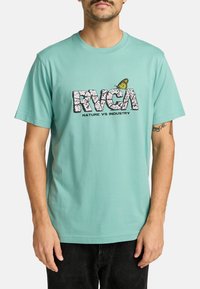 Camiseta de algodón en verde azulado claro con un logo impreso texturizado en negro "RVCA" que incluye el texto "NATURE VS INDUSTRY" y un pequeño gráfico de mariposa amarilla.