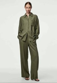 Ensemble en vert olive comprenant une chemise et un pantalon large en tissu doux. La chemise présente des boutons et une poche poitrine. Le modèle se tient debout dans des sandales plates.