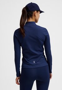 Marinblå långärmad idrottströja i elastiskt material, med hög krage och diskret logotypdetalj på baksidan nära fållen.