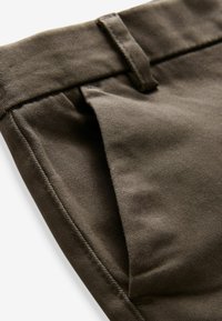 Tissu vert olive avec une texture lisse, présentant une passant de ceinture, une poche zippée et des détails de couture précis. Conception ajustée adaptée pour une tenue décontractée.
