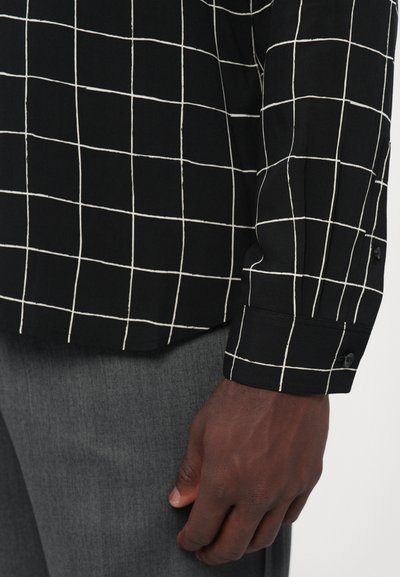 Zwarte shirt met wit ruitpatroon, zachte stof, lange mouwen, met knoopmanchetten, gecombineerd met grijze broek. Gedetailleerde stiksels zichtbaar.