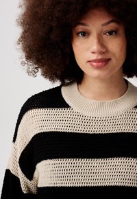 Jong persoon met krullend haar, gekleed in een zwart-beige gestreept gebreid vest, kijkt recht vooruit met een neutrale uitdrukking op een effen achtergrond.