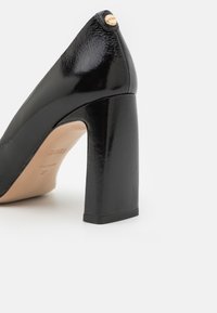 Escarpin à talon haut en cuir verni noir avec talon bloc épais et petit détail de logo rond doré à l'arrière.