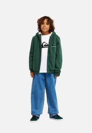 Boy with curly hair wearing a green zip-up hoodie, white t-shirt with black logo, loose blue jeans, and black and green sneakers standing.