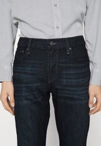 Jeans din denim închis cu închidere prin buton, design cu cinci buzunare și decolorare subtilă. Poartă o cămașă cu buton în gri deschis.