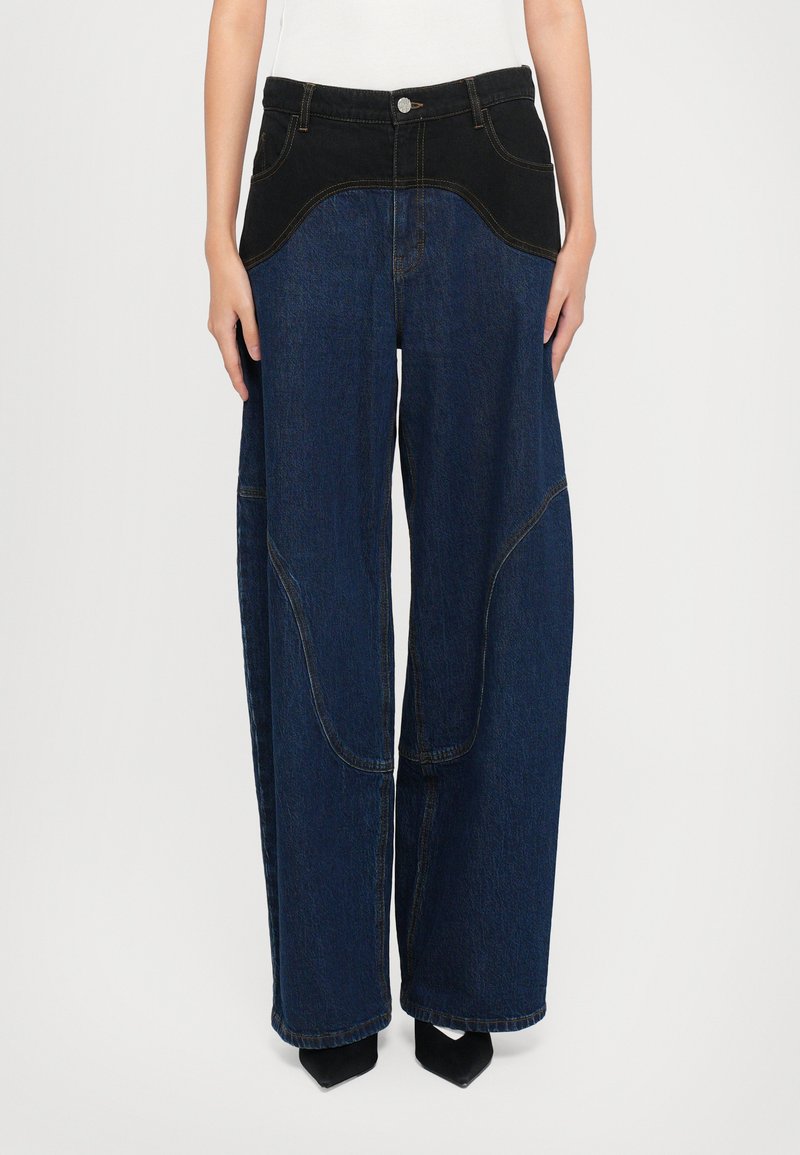 Laiad säärised teksad, millel on tumesinine denimkangas ja must ühtlane paneel vööl, nähtavate õmblustega ja kerge läikega.