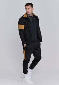 Black athletic tracksuit with gold accents, featuring a zip-up jacket and tapered pants. Worn with white sneakers, showcasing a fitted design.